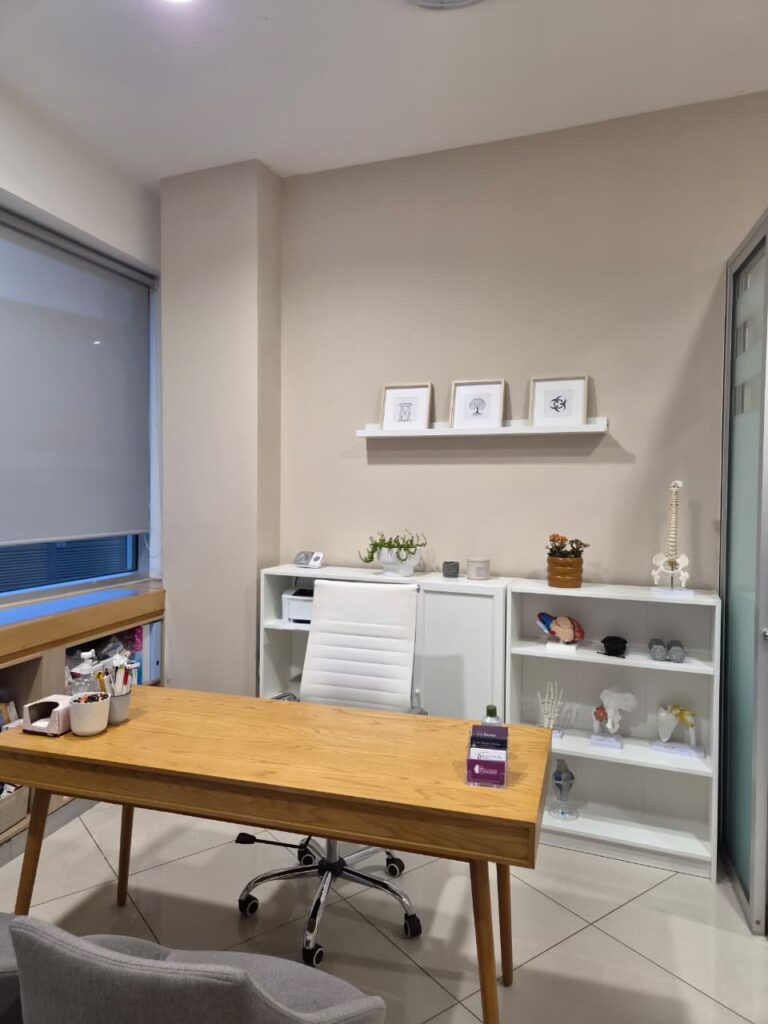 A modern office or clinic: wooden desk with white rolling chair, glass shelves with models and plants in a neutral-color room.